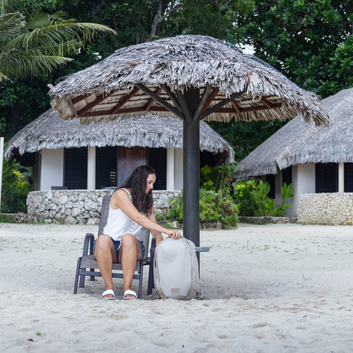 A woman sits on a beach chair under a thatched beach umbrella on a sandy beach, with tropical palm trees and thatched bungalows in the background.